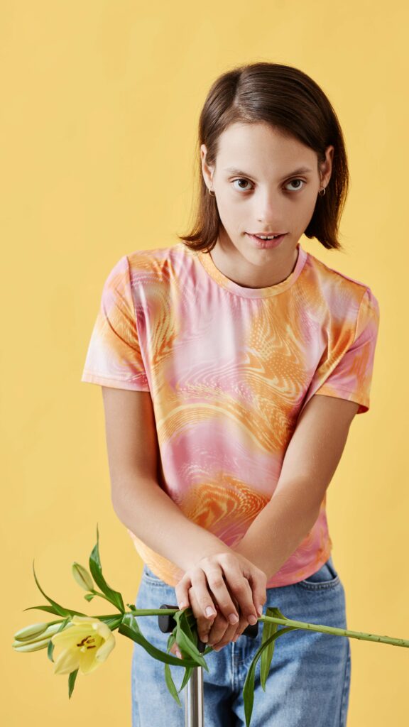 Retrato vertical de una niña joven con parálisis cerebral sosteniendo una flor de lirio y mirando a la cámara, de pie contra un fondo amarillo.